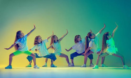Synchronous movements. Group of children, little girls in sportive casual style clothes dancing in choreography class isolated on green background in neon light. Concept of music, fashion, art