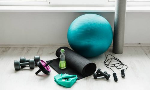 Group of different exercising equipment on white home gym floor. Fitness ball, round foam roller, resistance exercise latex band, jumping rope, dumbbells, yoga mat. Fit lifestyle concept.